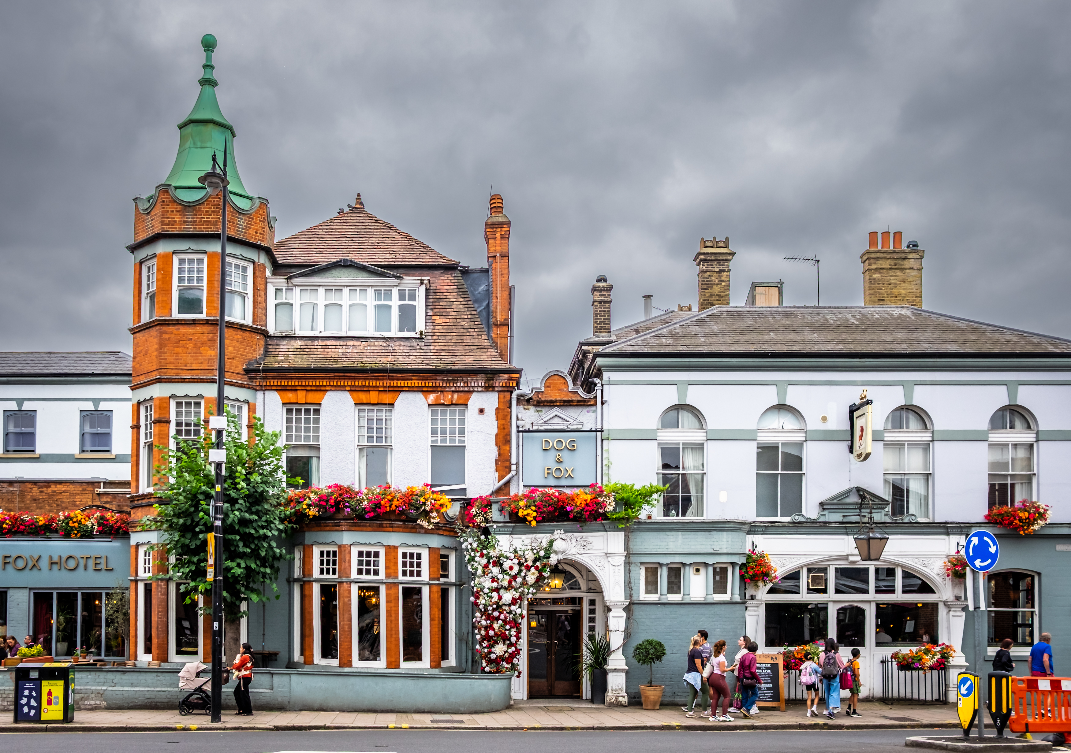 The Dog and Fox pub and hotel in Wimbledon Village, with floral decorations and people outside beneath a cloudy sky.